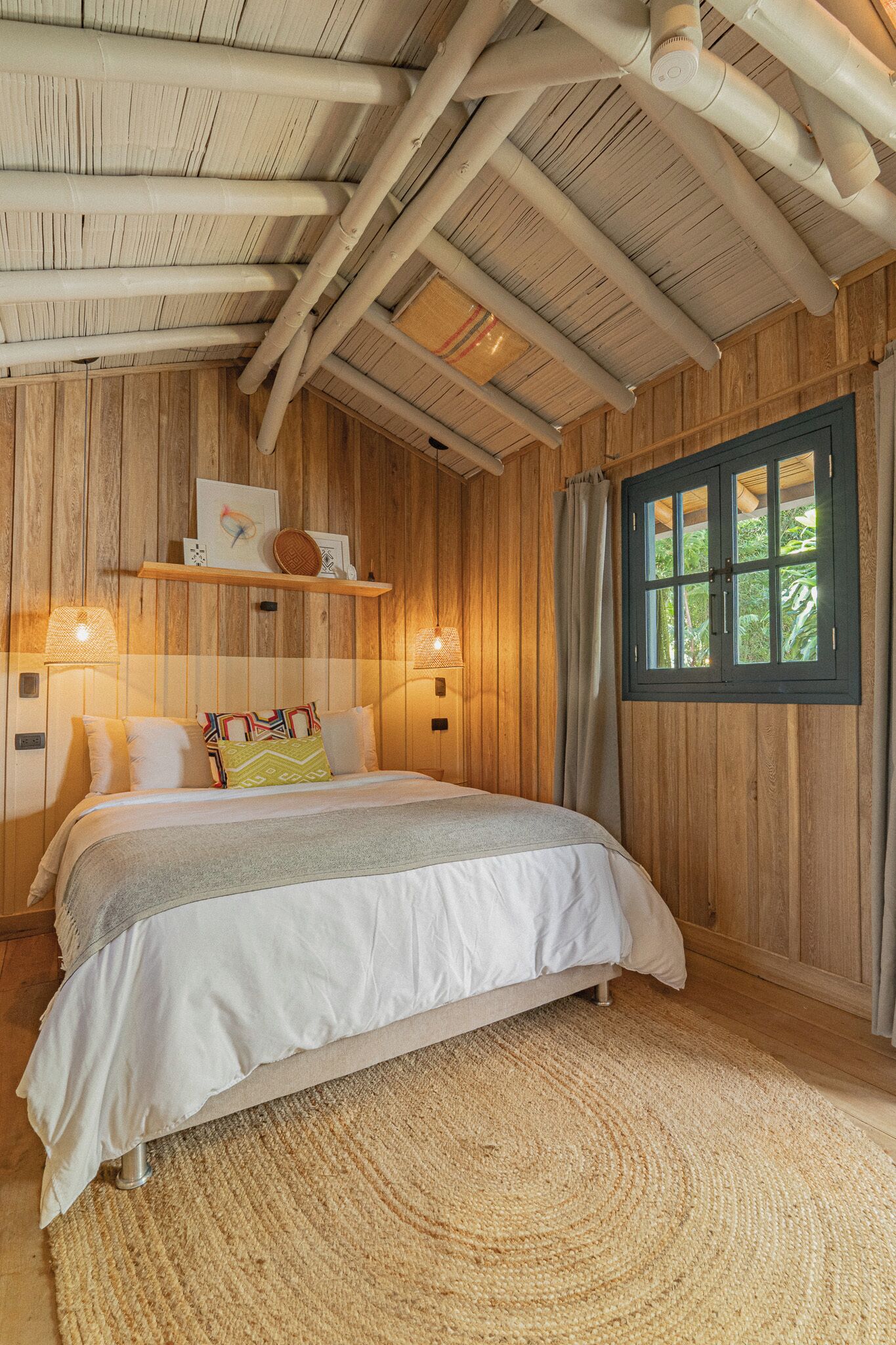 A wooden-paneled room with a bed, wall-mounted lamps, a window with curtains, and a shelf with decorative items.