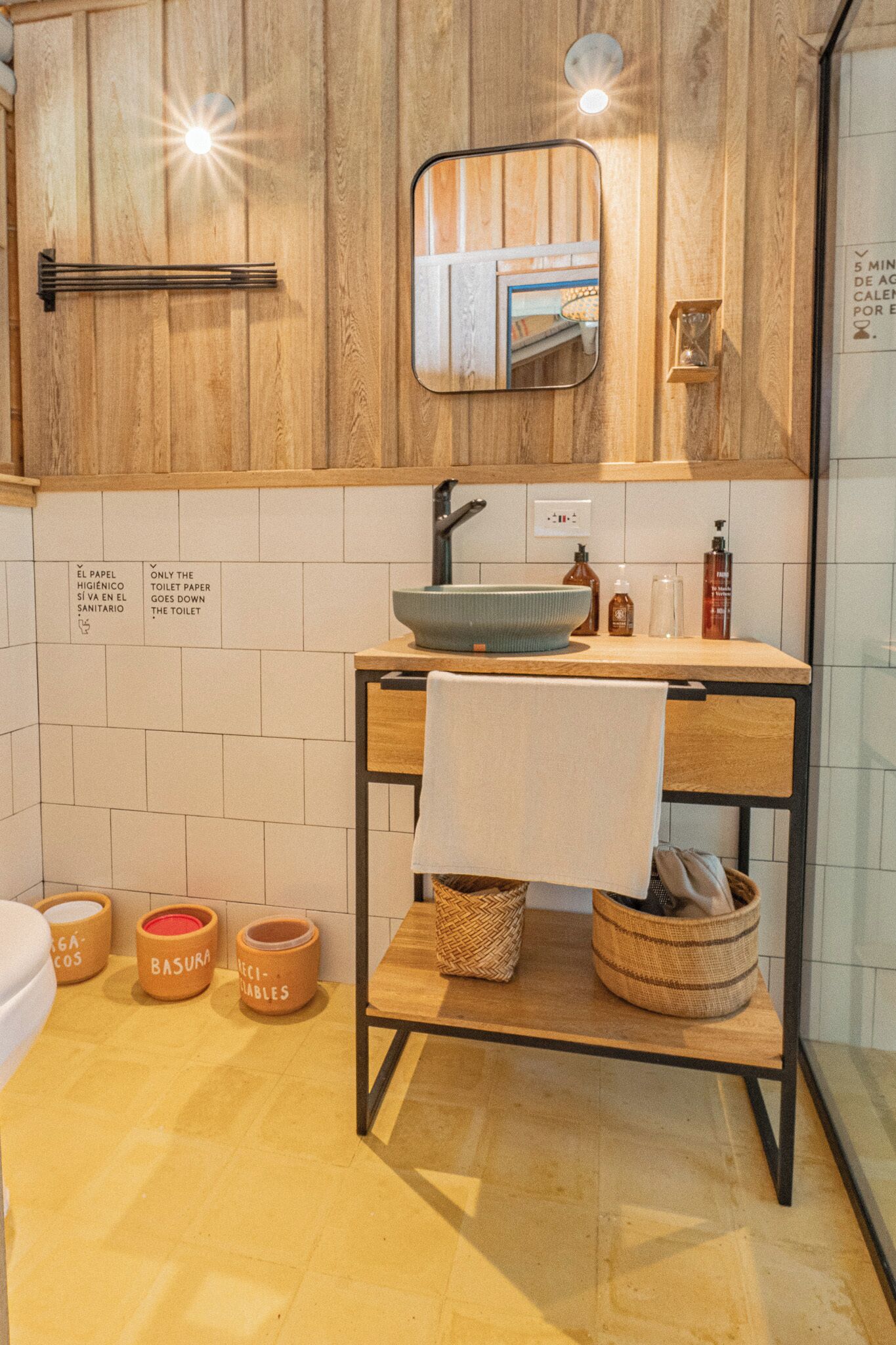 A bathroom with a wooden vanity, a mirror, a sink, and a towel hanging on the wall. There are labeled containers on the floor.