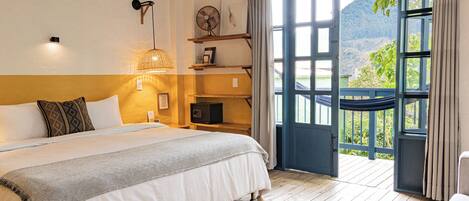 A bedroom with a bed, bedside lamps, a fan, and a view of a balcony with a mountain.