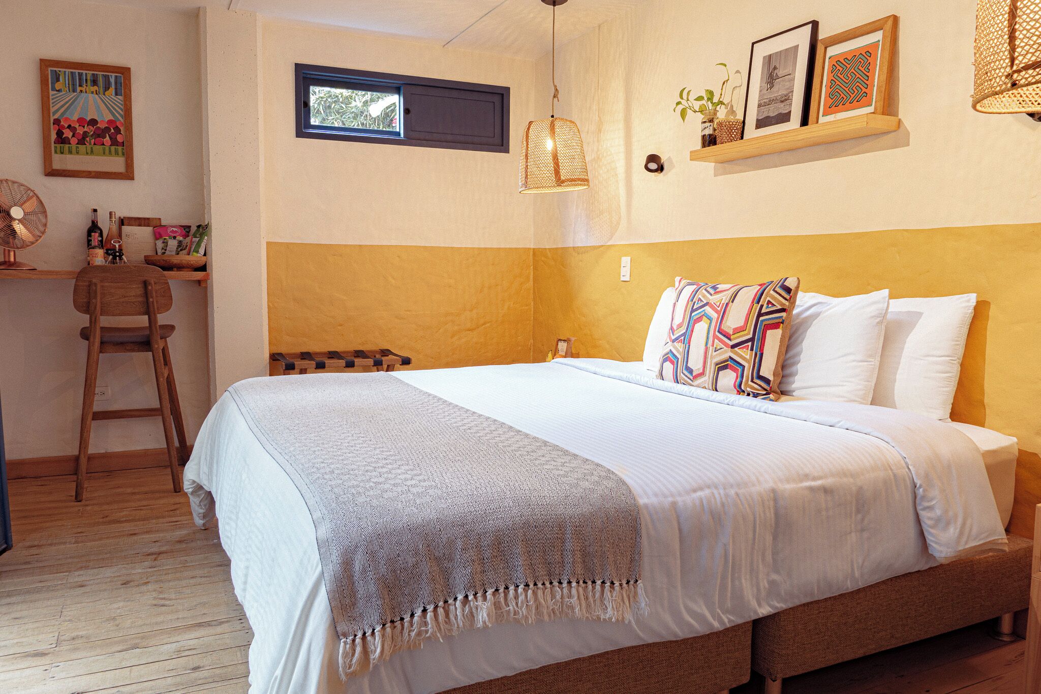 A neatly made bed with a patterned throw, a wooden chair, a desk with a fan, and a small window.