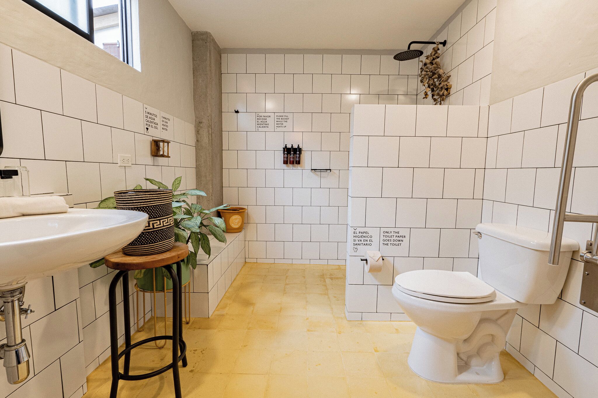 Une salle de bain équipée d’un lavabo, de toilettes et d’une zone de douche, avec des carreaux blancs et un tabouret en bois.