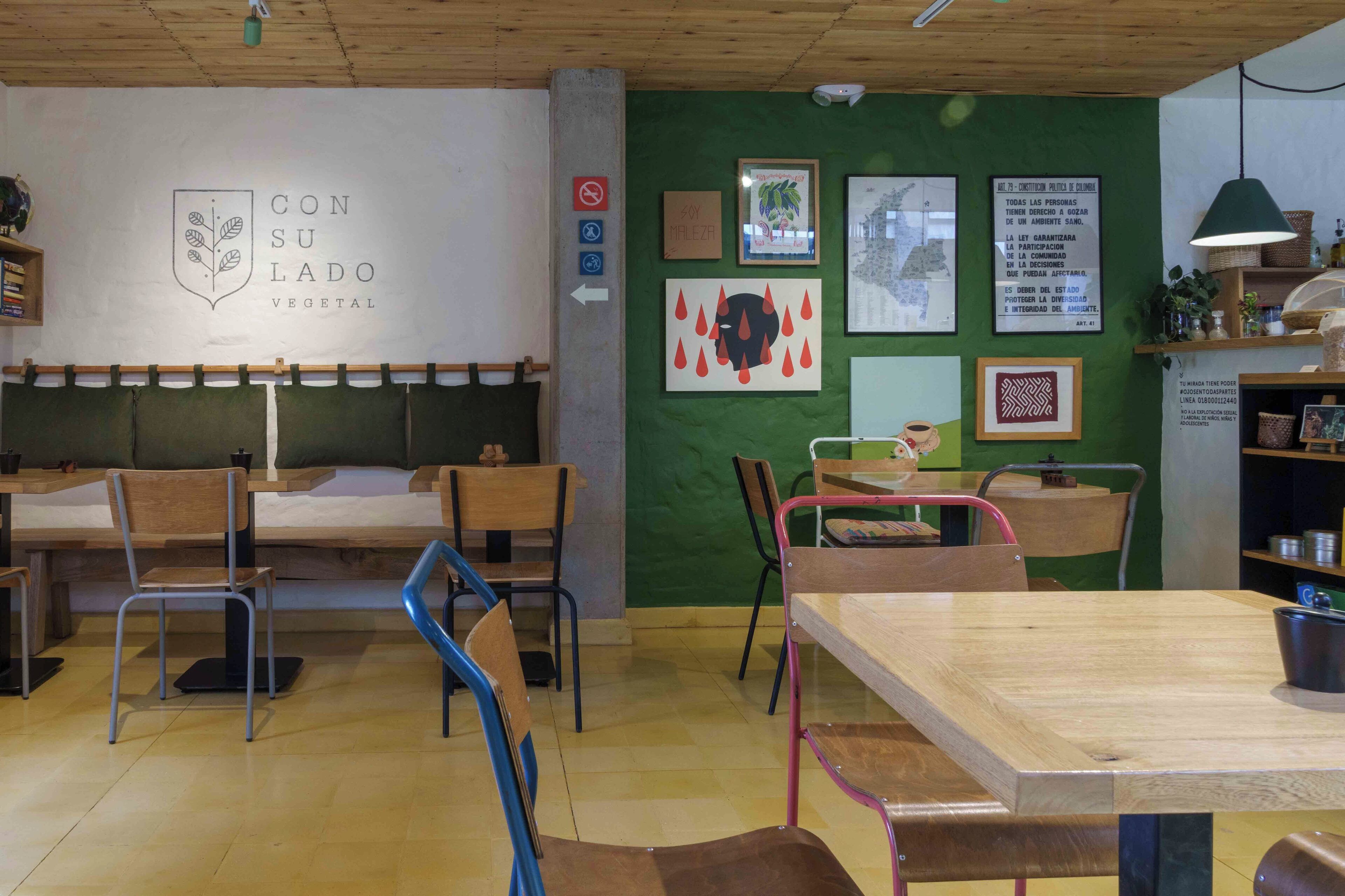 A cozy café with wooden furniture, a green wall with various framed pictures, and a sign indicating a vegetarian option.