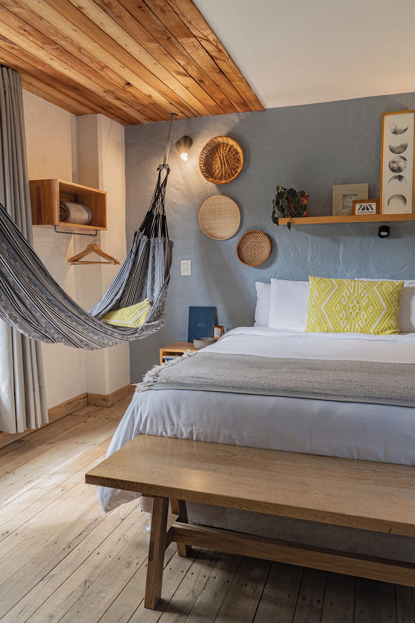 A bedroom with a bed, a hammock, a wooden bench, and a wall with decorative items.