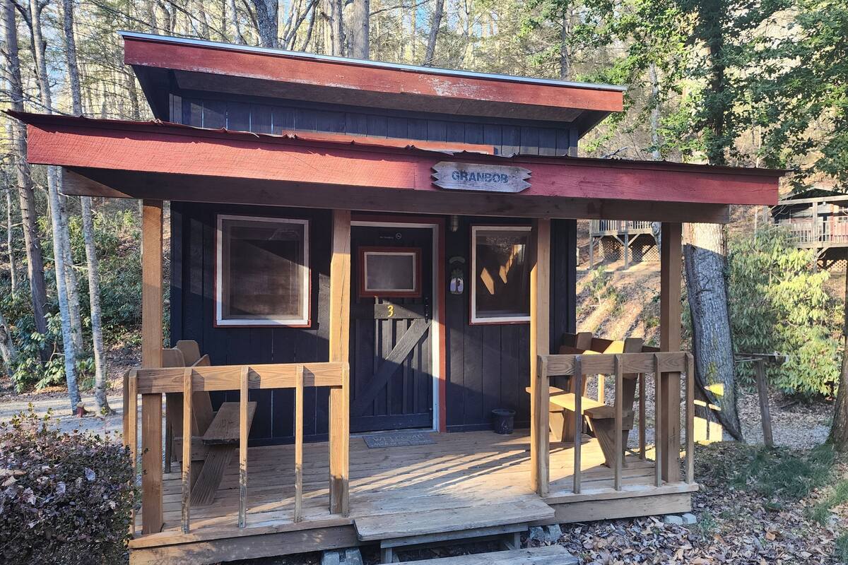 A wooden cabin with a red roof, labeled "GRANBOR," featuring a porch with wooden benches and a small staircase leading to the entrance.