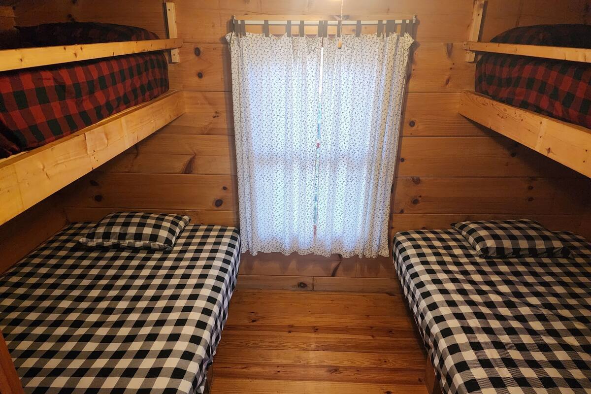 A cabin with two bunk beds, each with a checkered bedspread, a wooden floor, and a curtain with a pattern.