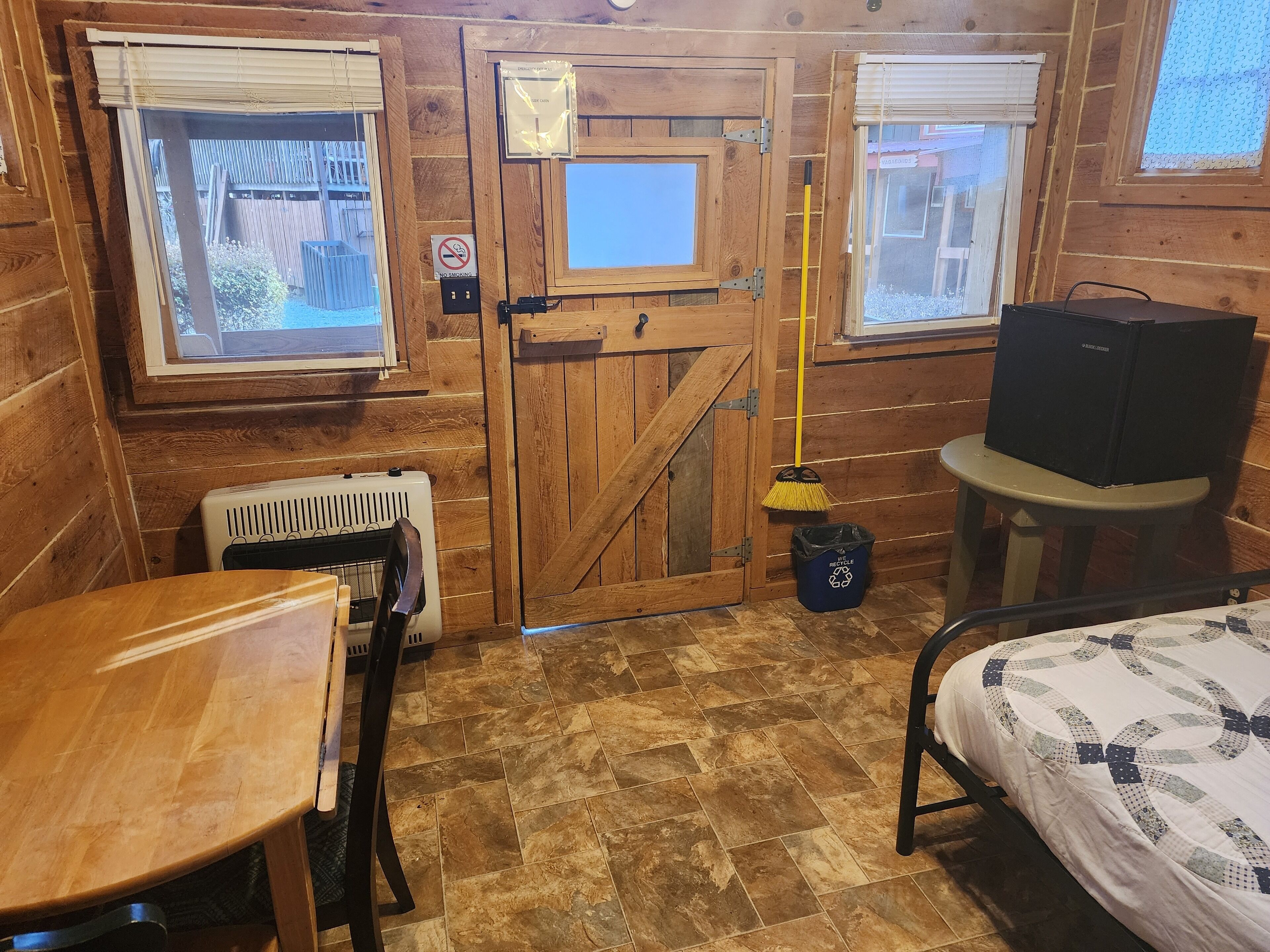A wooden cabin room with a bed, a small table, a radiator, a broom, and a trash bin.