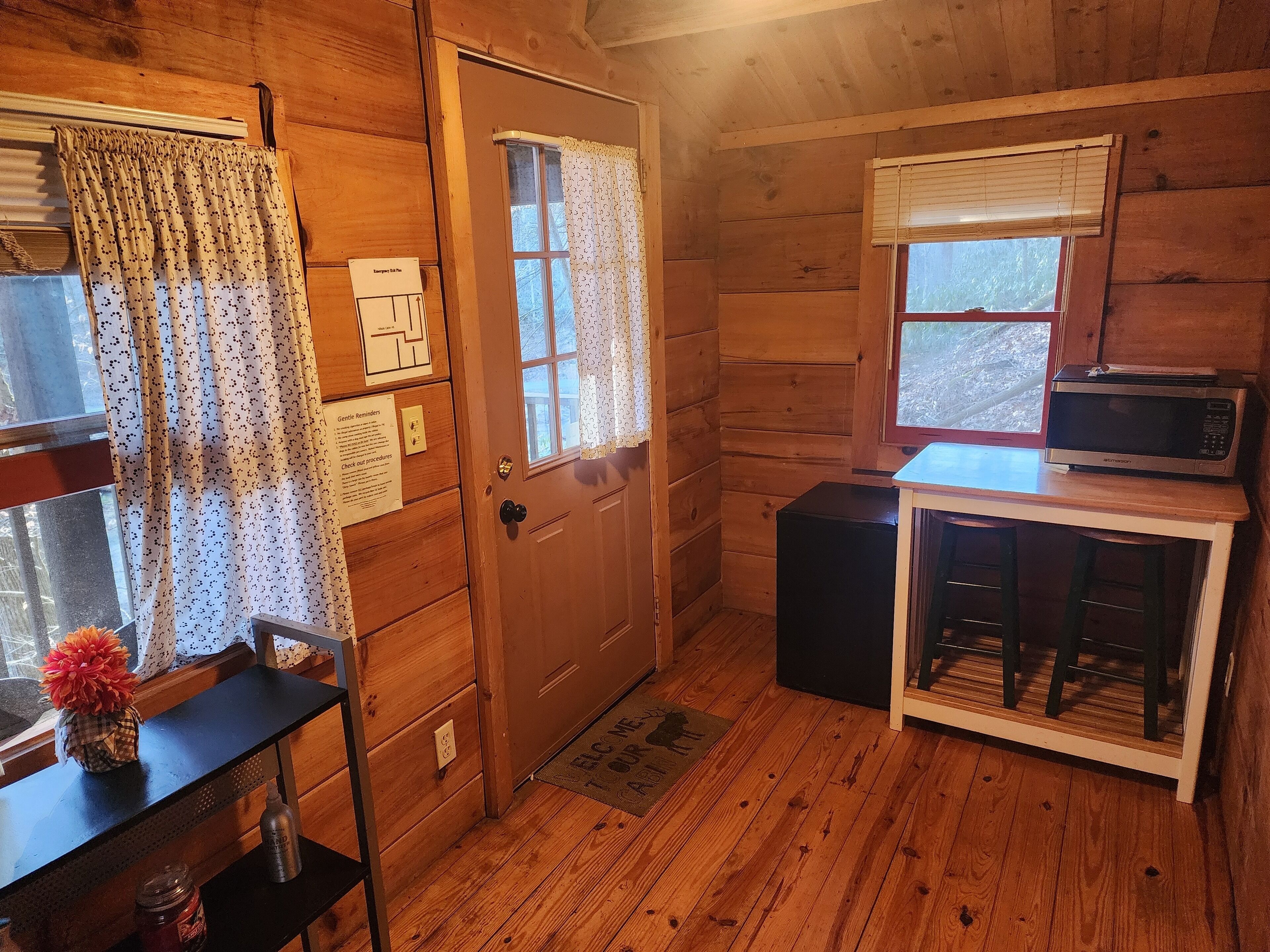 L’intérieur d’une cabane en bois, avec une porte, une fenêtre avec des rideaux, un four à micro-ondes posé sur une petite table et une bouteille sur une étagère.