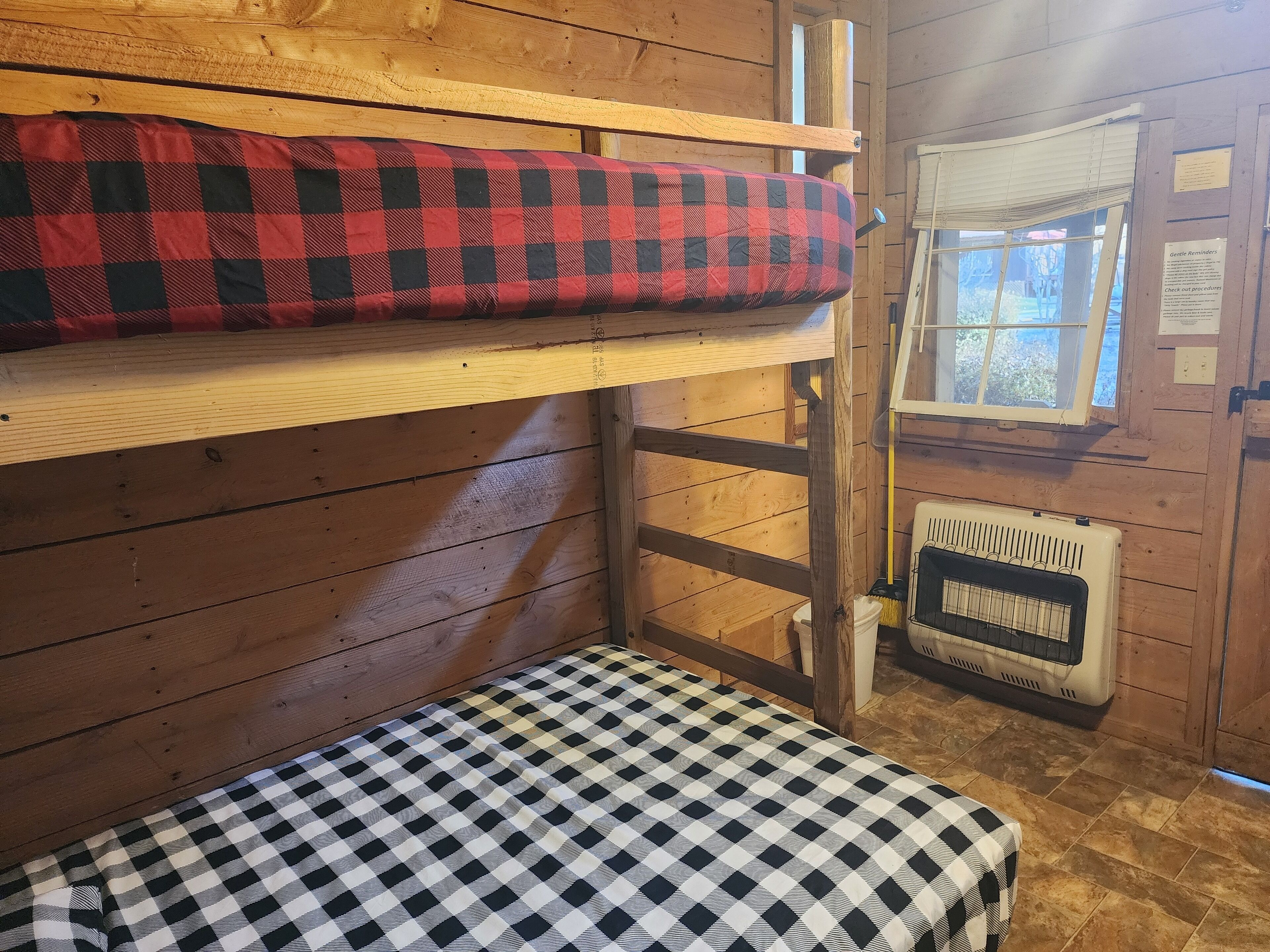 A bunk bed with a red and black checkered blanket and a black and white checkered blanket. There is a window with blinds and a radiator below the window.