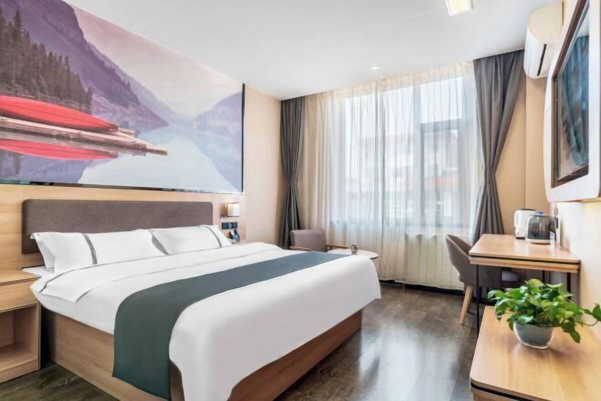 A hotel room with a bed, a desk, a chair, a plant, and a painting on the wall.