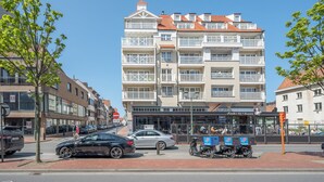 A multi-story building with balconies, a restaurant at the ground level, and parked cars and scooters on the street. - Apartment in Knokke Near Lippenslaan (Knokke-Heist)