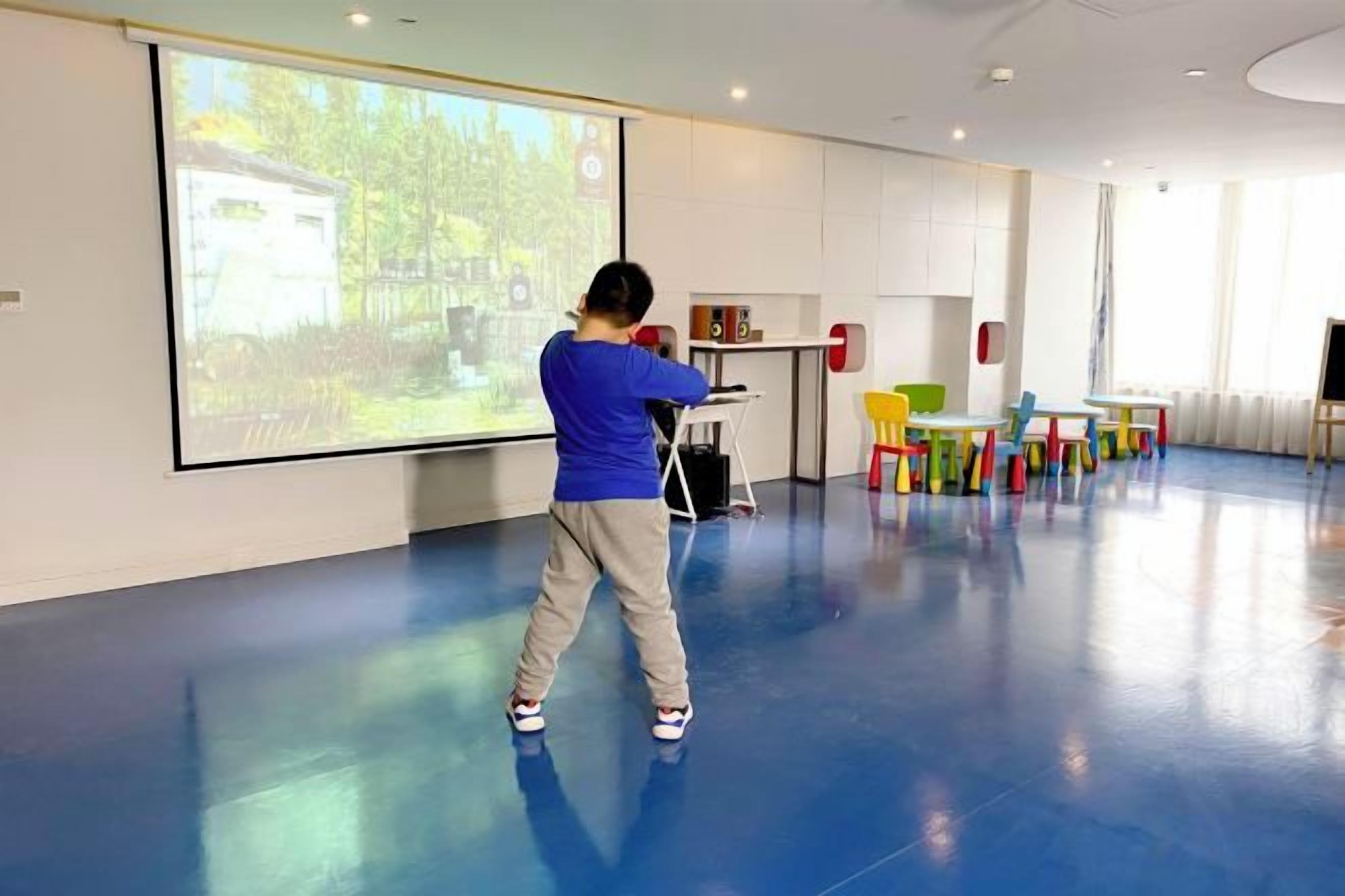 A child playing with a large screen in a room with a blue floor, a projector, and colorful chairs.