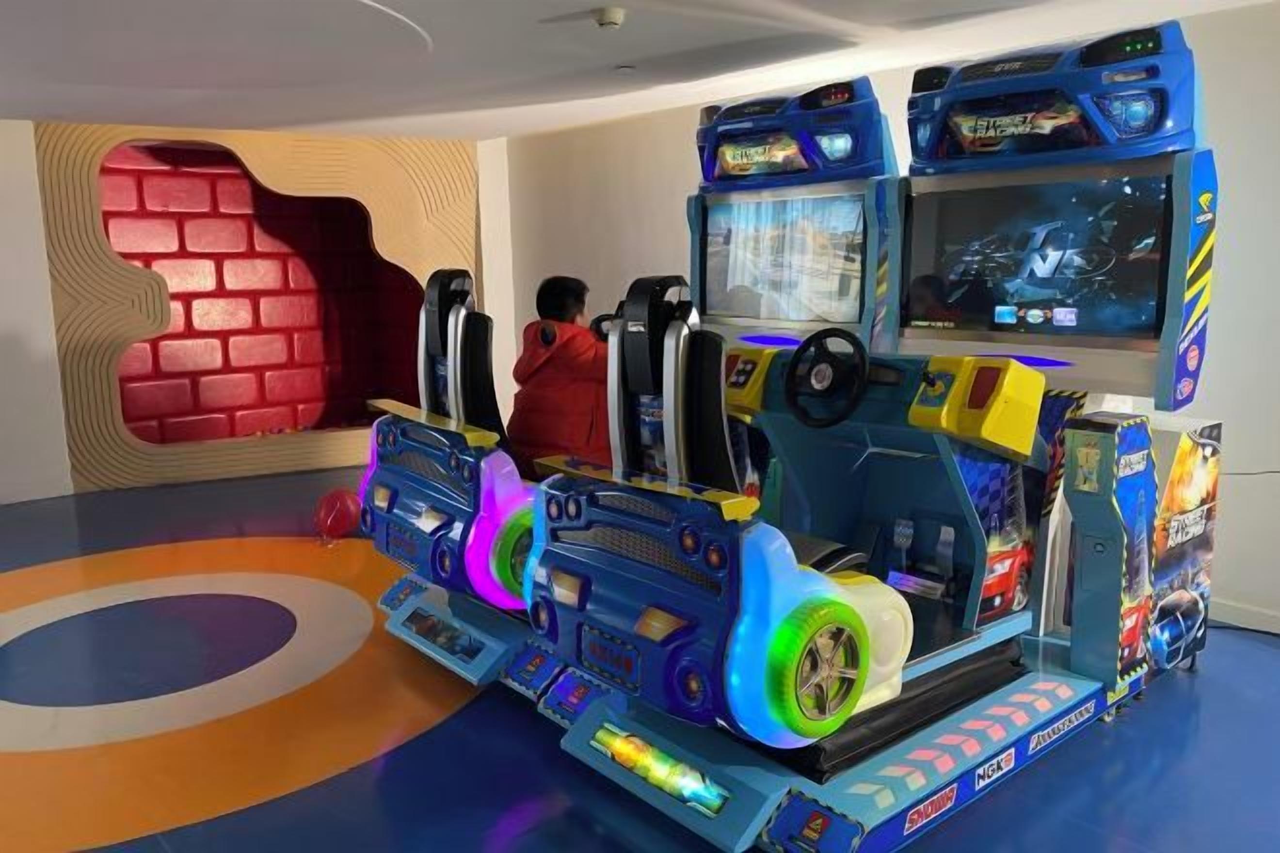 Une salle d’arcade proposant plusieurs jeux de course, avec un mur en briques rouges et un sol aux motifs colorés.