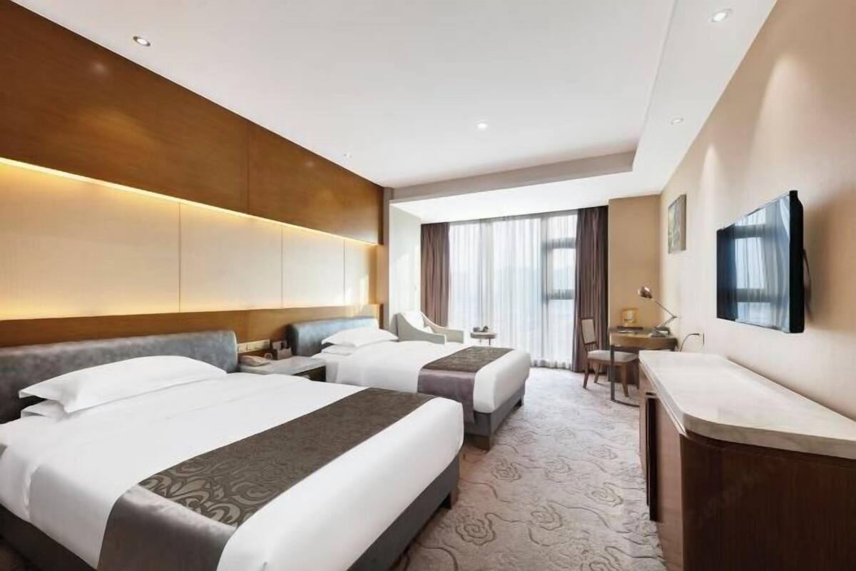 A hotel room with two beds, a desk, a chair, a TV, and a large window.