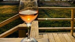A glass of wine on a wooden deck with a view of the ocean and a sunset in the background. - Designer Waterfront Bay House w/ Direct Access, Deck & Fire Pit (Shirley)