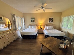 A hotel room with two beds, a desk, a dresser, a ceiling fan, and a window with blinds.