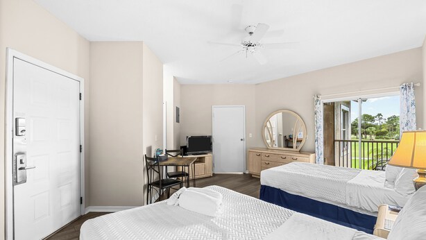Quarto de hotel com duas camas, uma escrivaninha com computador, um ventilador de teto e varanda com vista.