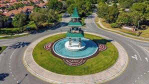 Uma rotatória com uma fonte central, rodeada por vegetação e céu claro.