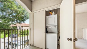 Uma lavanderia com máquina de lavar e secadora, uma porta e vista para a rua com carros estacionados.