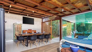 A modern dining area with a wooden table and chairs, a large window, and a view of a pool and greenery outside. - Bz70 Casa Piscina 650m da Rua das Pedras (Buzios)