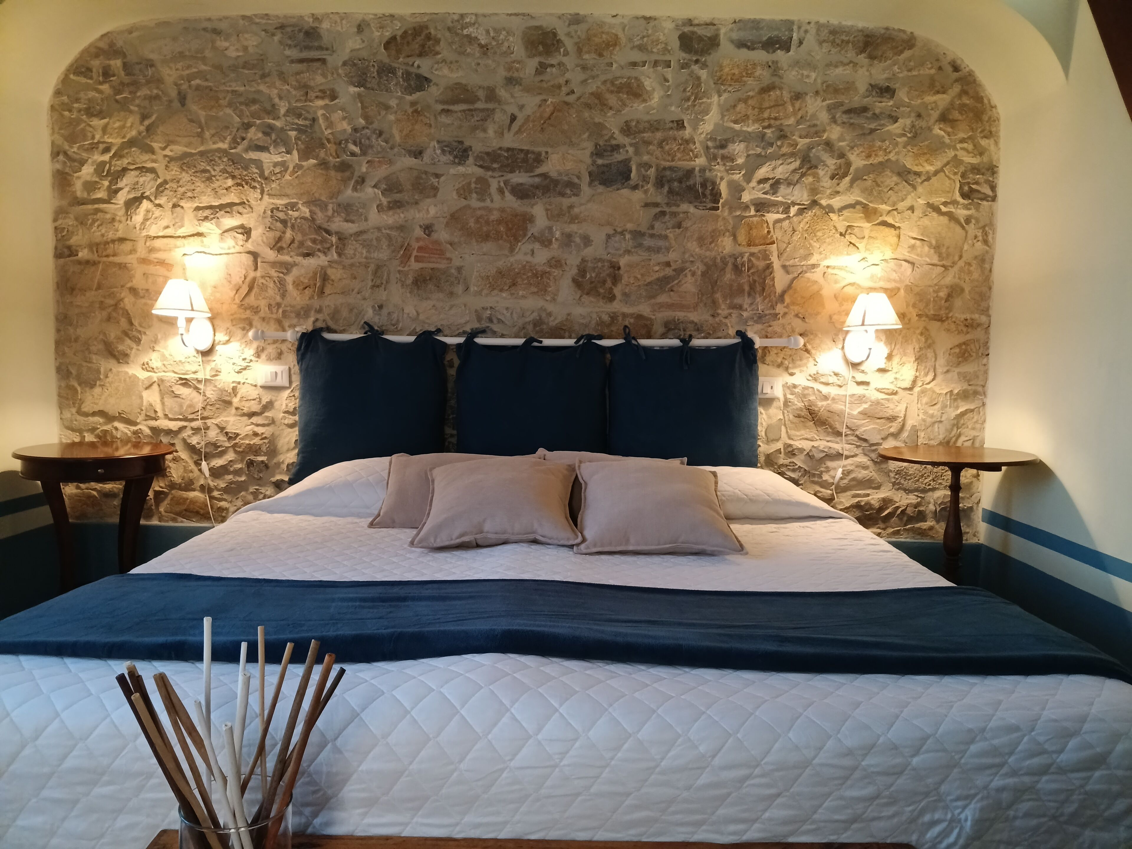 A bedroom with a stone wall, a bed with a quilted cover, two bedside tables, and wall-mounted lamps.