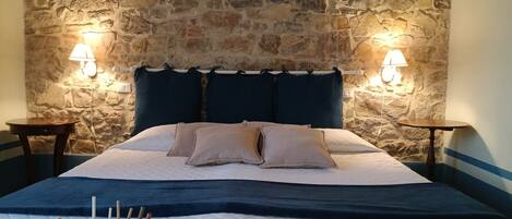 A bedroom with a stone wall, a bed with a quilted cover, two bedside tables, and wall-mounted lamps.