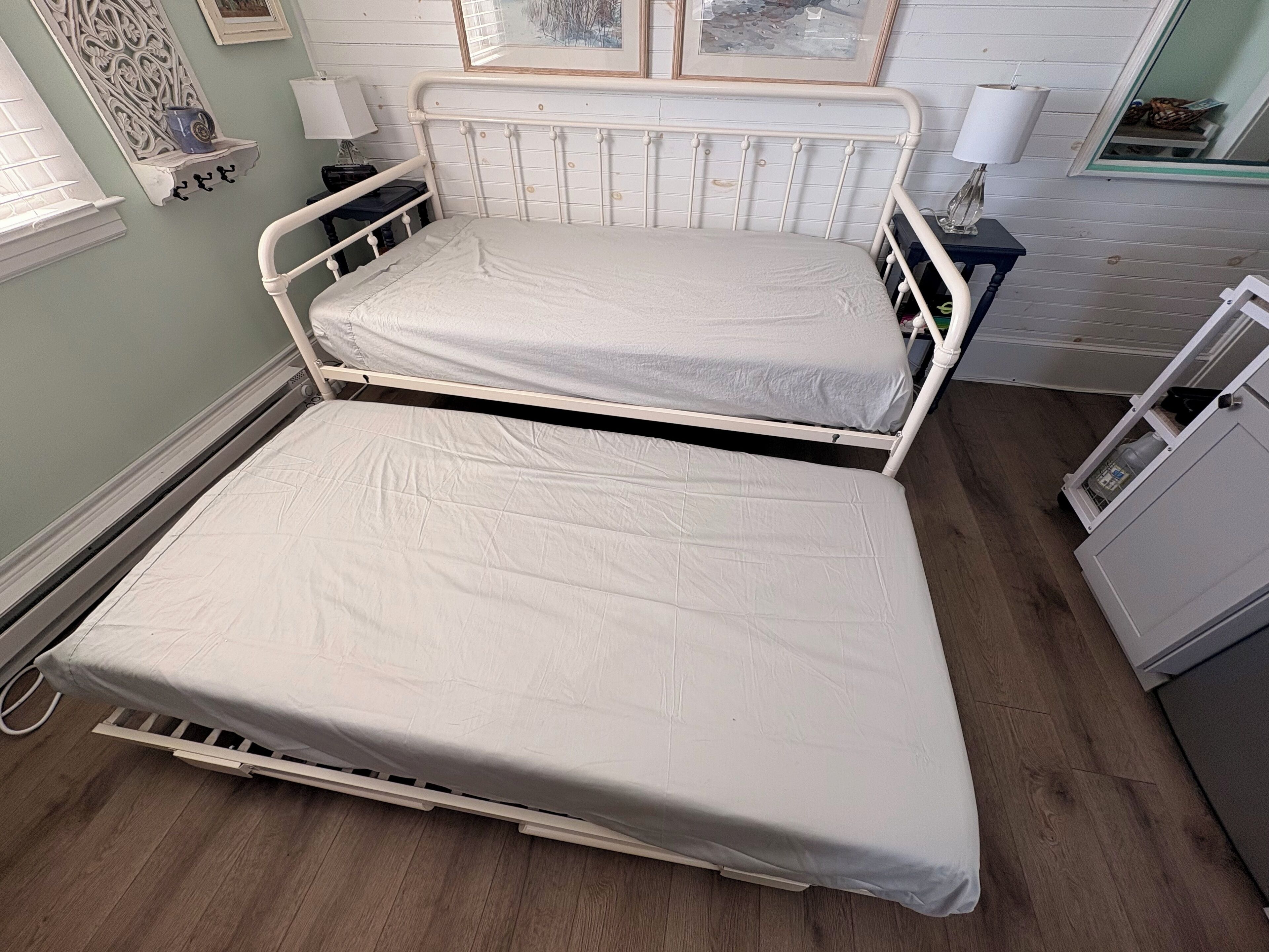 A twin bed with a white mattress in a room with light green walls, a wooden floor, and a small bedside table with a lamp.