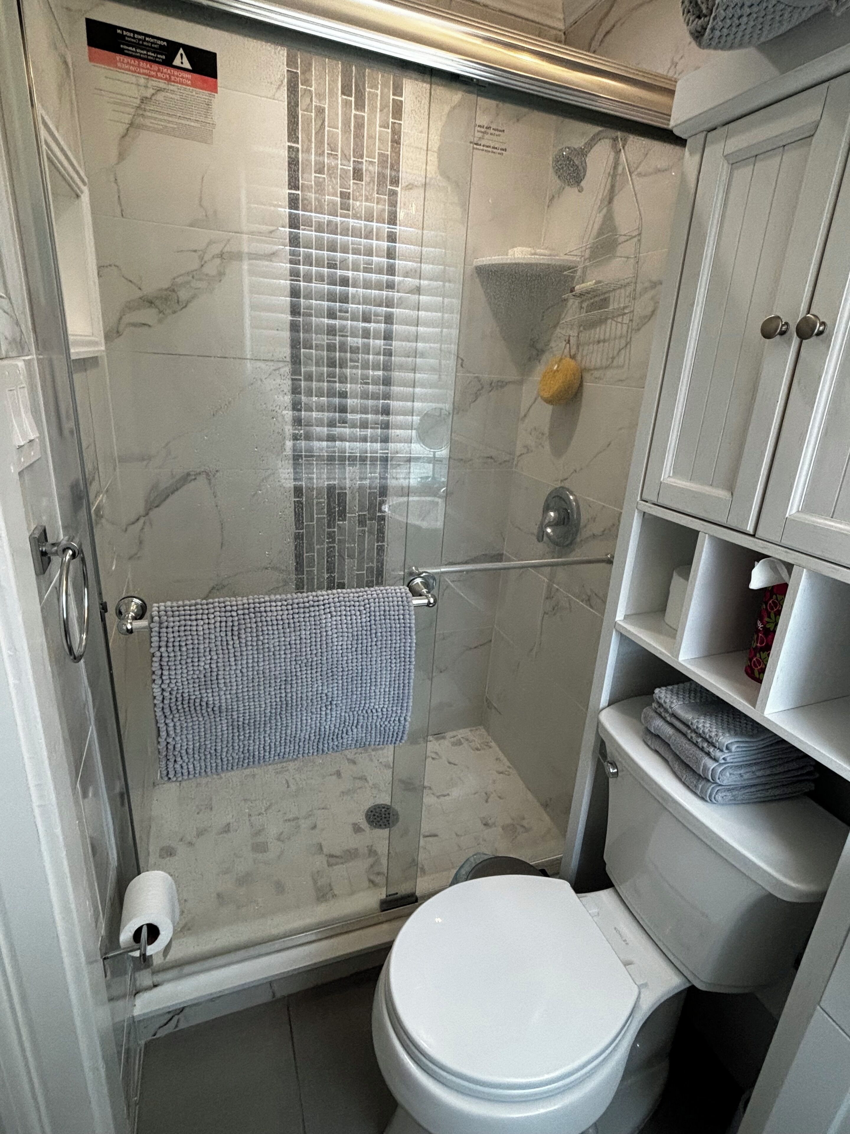 A compact bathroom with a glass-enclosed shower, a toilet, and a small shelf with towels.