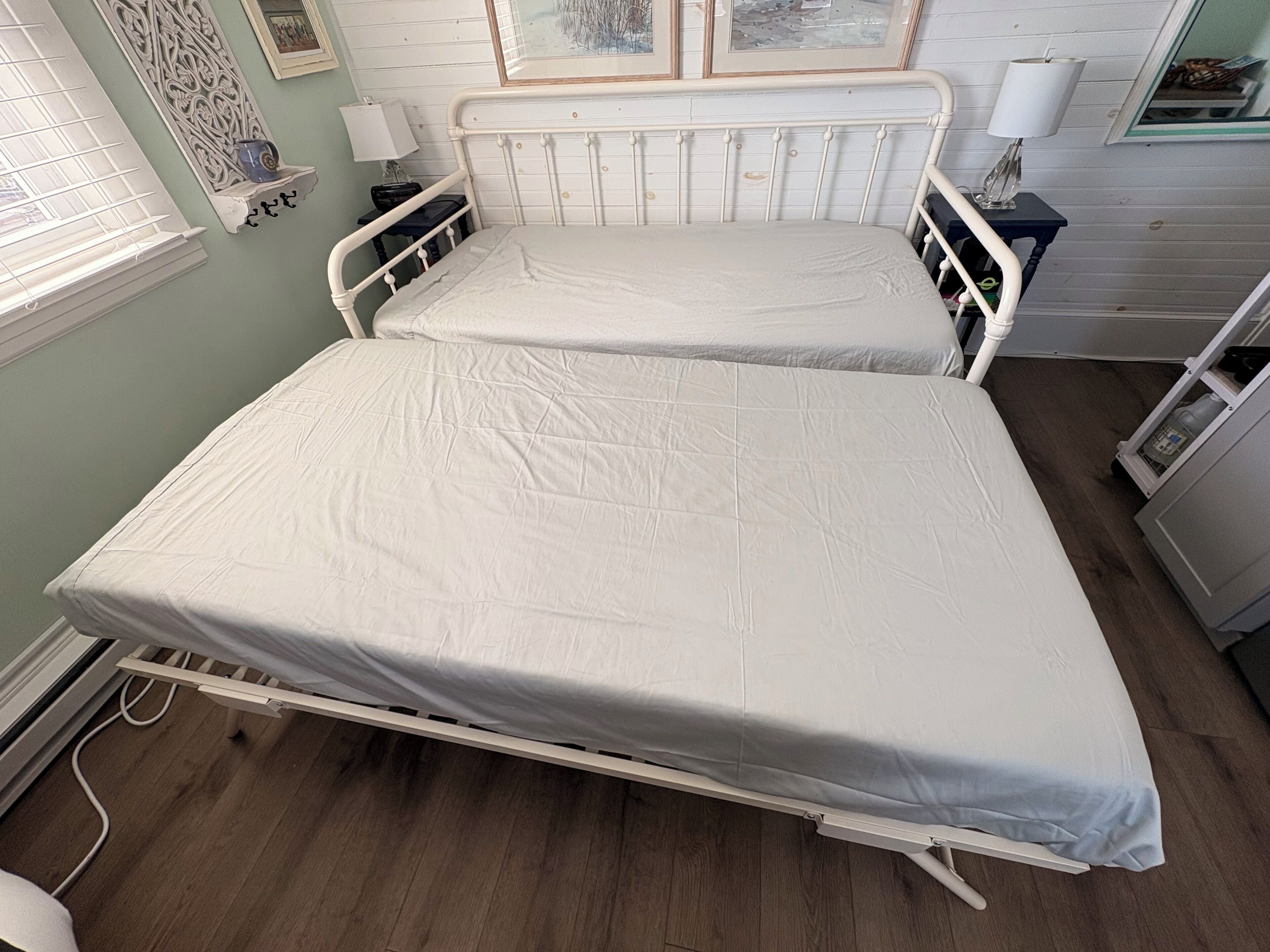 A double bed with a white mattress in a room with light green walls, a wooden floor, and a window with blinds.