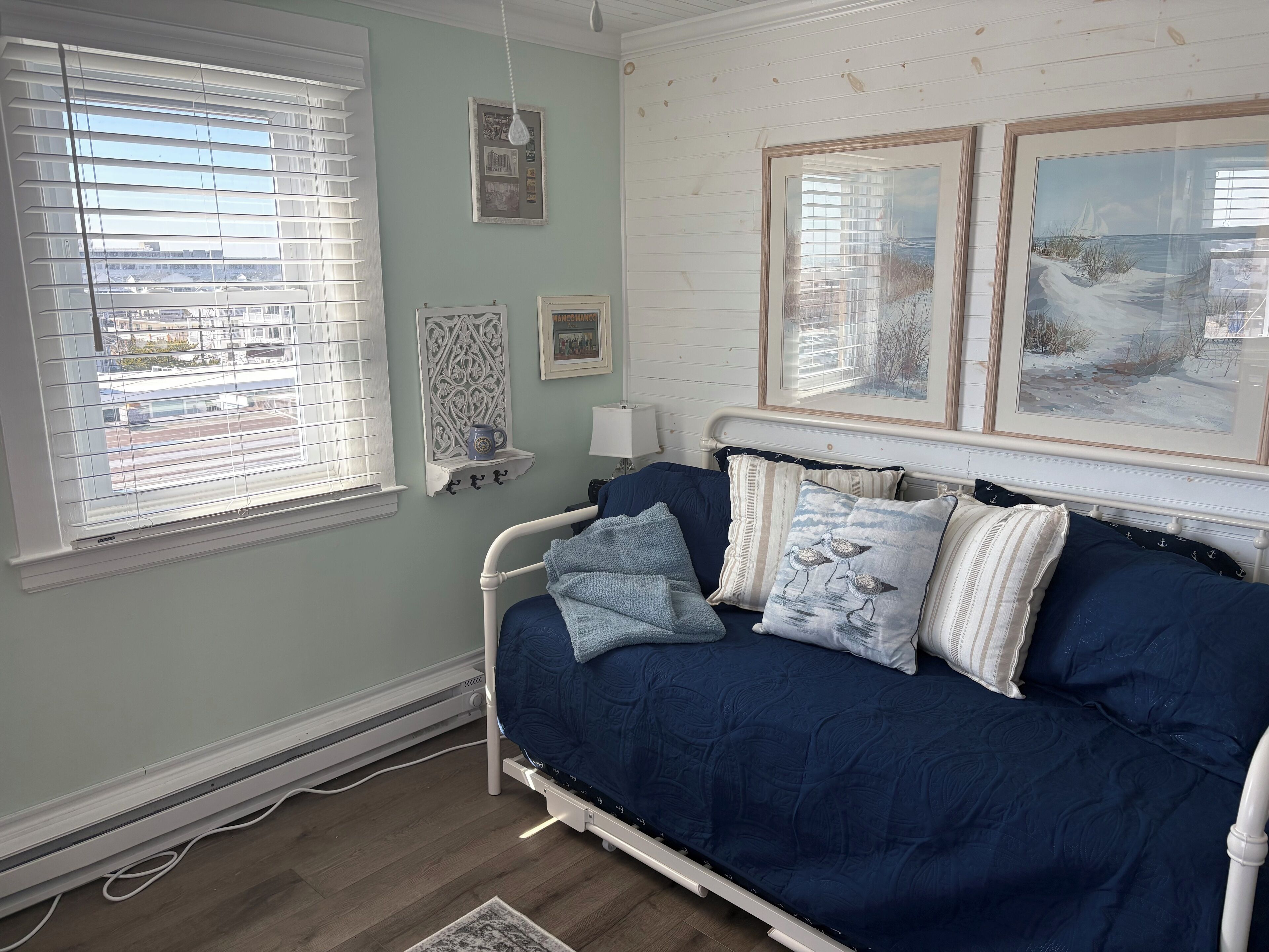A room with a bed, a window with blinds, a wall-mounted shelf with a mug, and framed pictures.