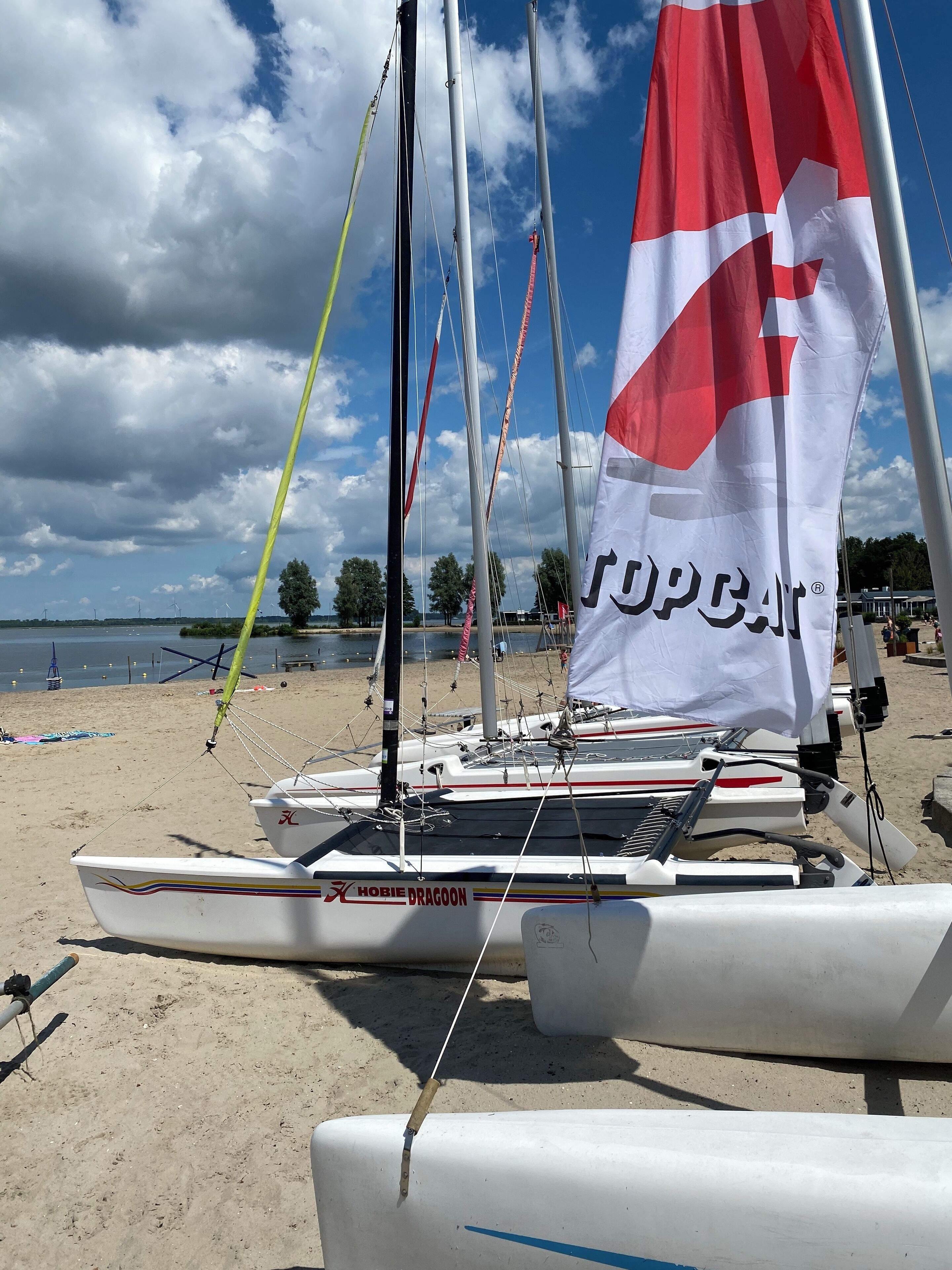 Un voilier baptisé « Hobie Dragoon » était amarré sur une plage de sable, un drapeau flottant au vent, sous un ciel partiellement nuageux.
