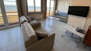 Uma sala de estar moderna com sofá, mesa de centro e TV. Janelas amplas oferecem vista para a praia.