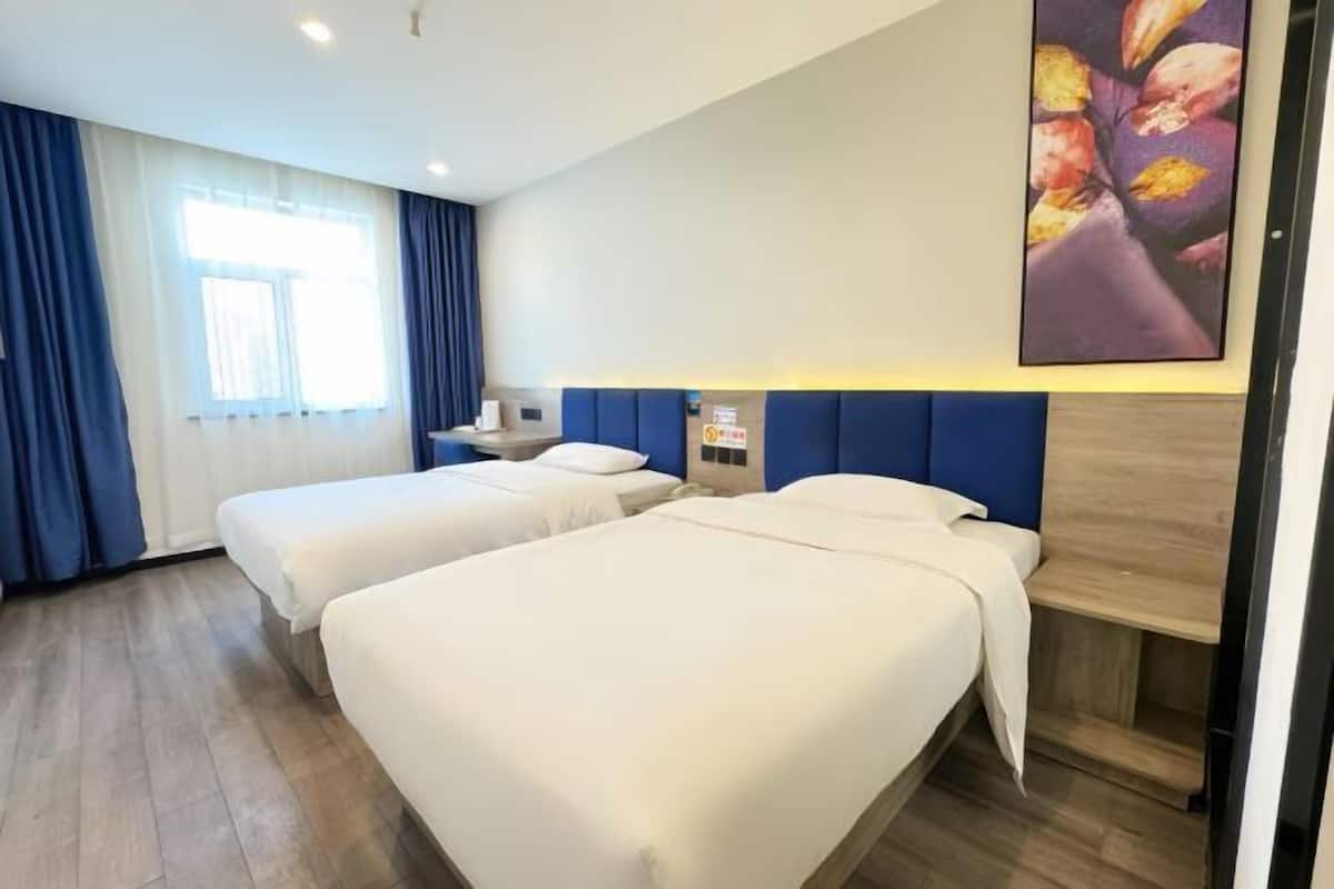 A hotel room with two beds, a large window with curtains, a wall-mounted artwork, and a wooden headboard.