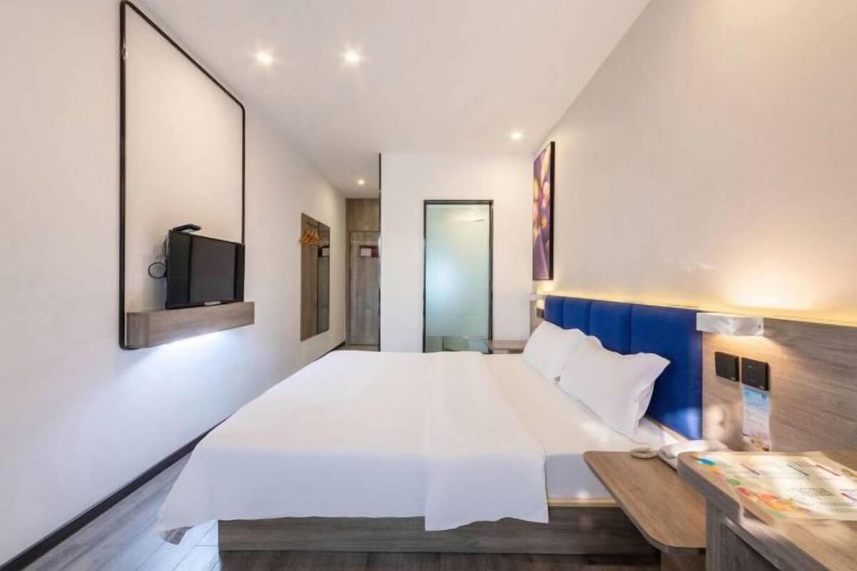 A modern hotel room with a large bed, a bedside table with a lamp, a flat-screen TV mounted on the wall, and a door leading to a bathroom.