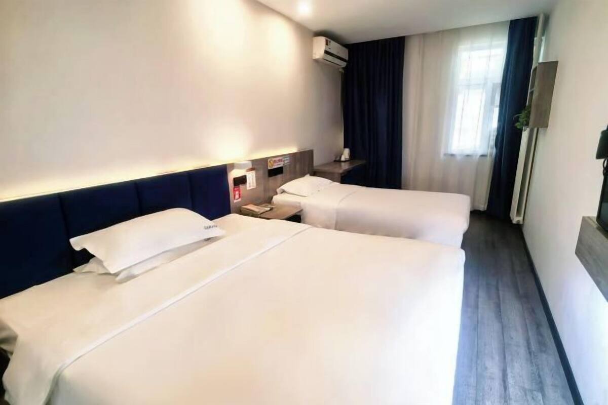 A hotel room with two beds, a bedside table, an air conditioning unit, a window with curtains, and a wall-mounted plant.