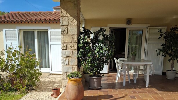 Un patio cubierto con un pilar de piedra, una planta en maceta, una mesa y sillas blancas, y un pequeño jardín con grava y plantas.