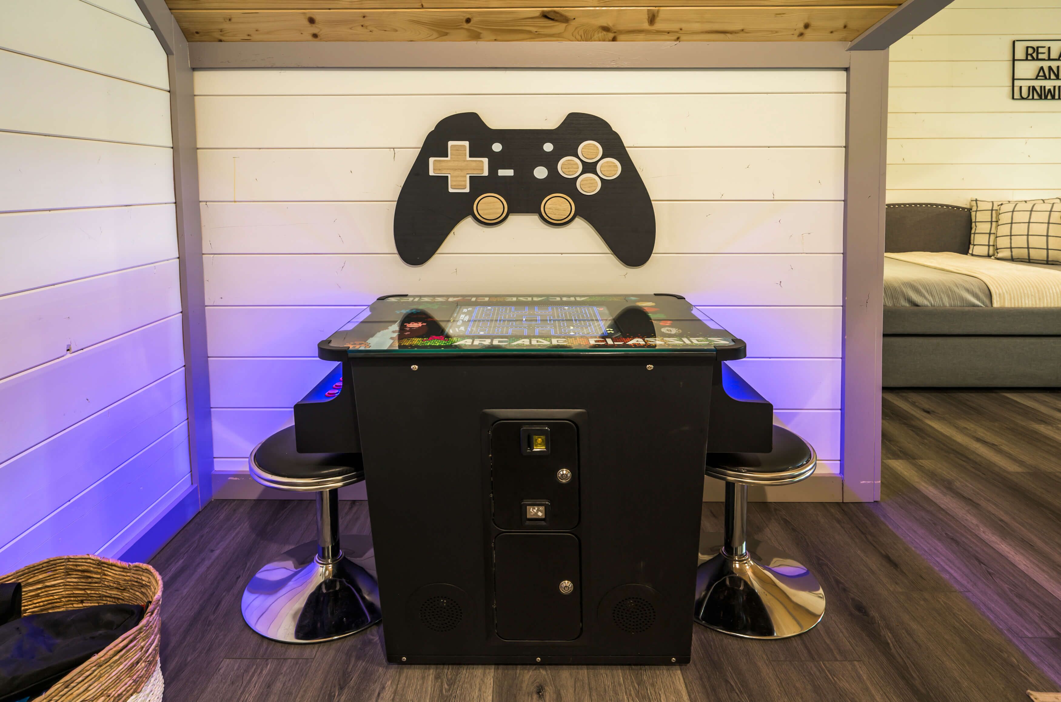 Une pièce avec un plafond en bois, une décoration murale représentant une manette de jeu vidéo, une table avec une console de jeux vidéo, deux tabourets de bar et un lit avec un oreiller à carreaux.