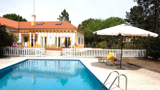 Una piscina con una cerca blanca, una sombrilla blanca y un edificio de paredes amarillas y blancas al fondo.