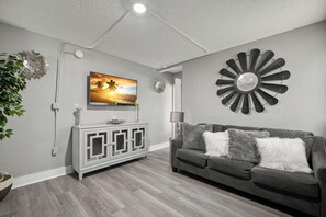 Ein Wohnzimmer mit einem an der Wand befestigten Flachbildfernseher, einer dekorativen Wanduhr, einem grauen Sofa mit zwei Kissen und einer weißen Kommode mit Glastüren.