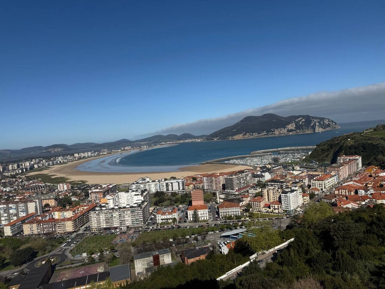 Un paisaje costero con edificios, una playa y montañas al fondo.