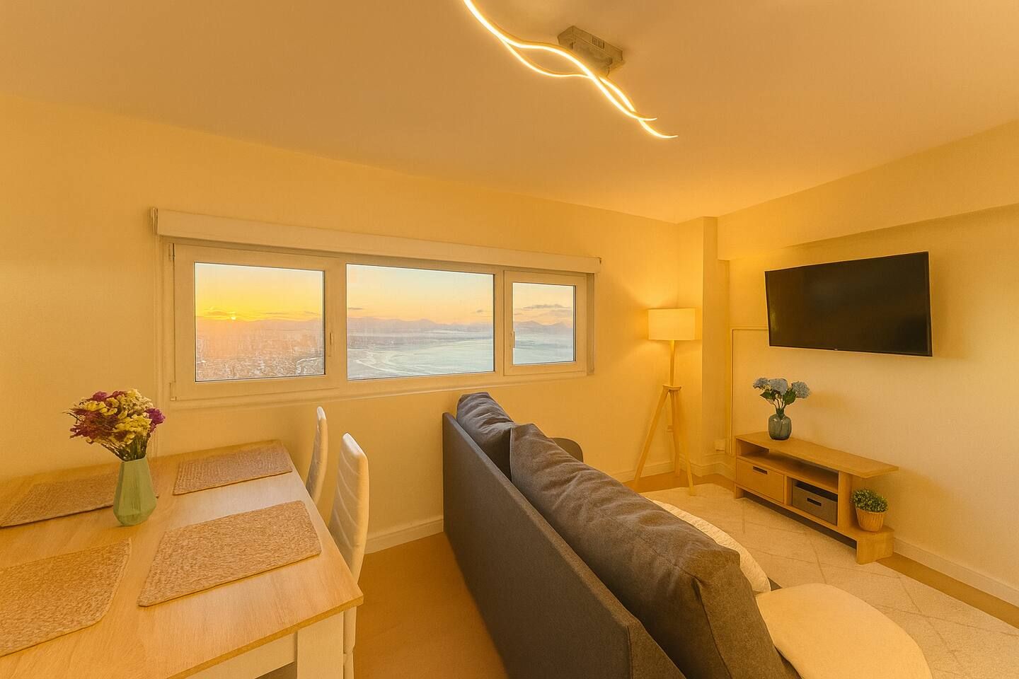 A room with a dining table, a sofa, a TV mounted on the wall, and a window with a view.