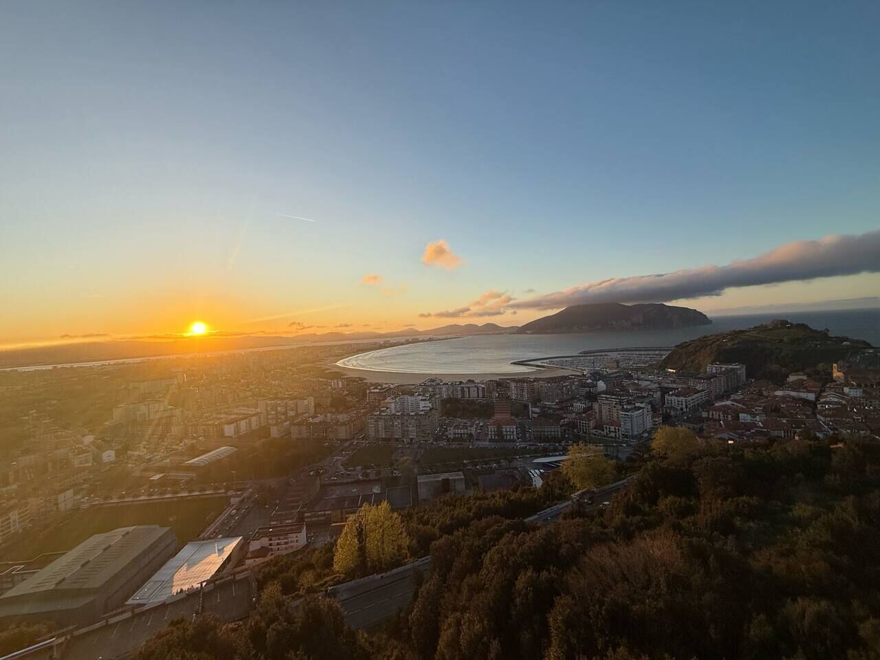 A coastal cityscape at sunset with a prominent hill and a body of water in the background.