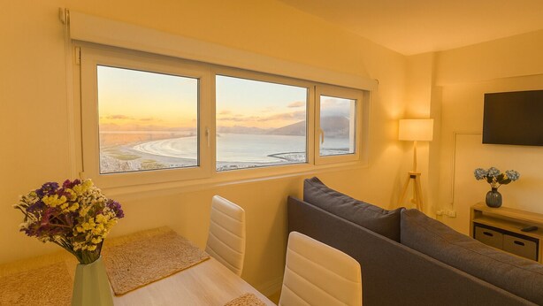 A room with a large window overlooking a beach, a sofa, a small table with a vase of flowers, and a TV on the wall.