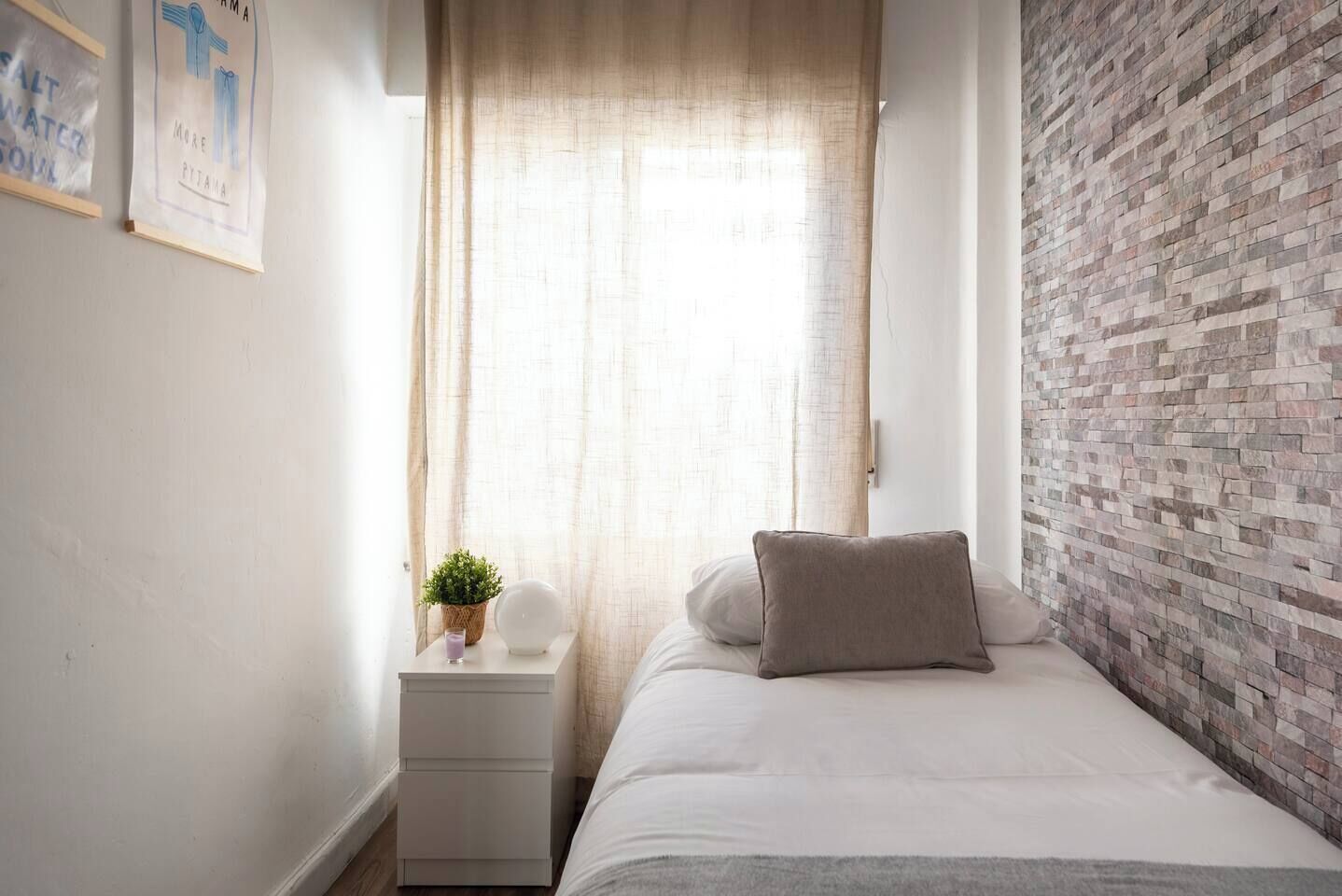 Ein Schlafzimmer mit einem Bett, einem Nachttisch, einer Zimmerpflanze, einer Wanduhr und einem Fenster mit Vorhängen.