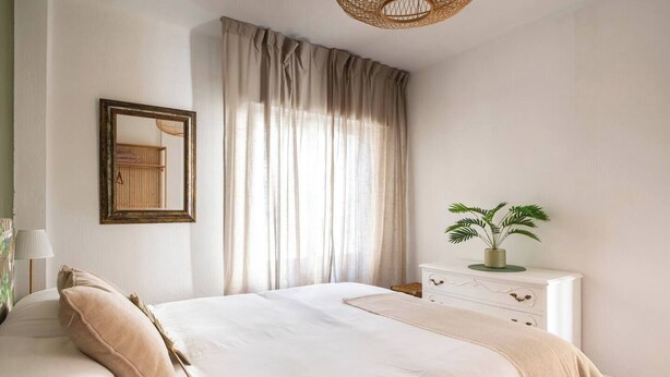 Quarto com uma cama grande, mesa de cabeceira com planta, espelho e cortinas leves.