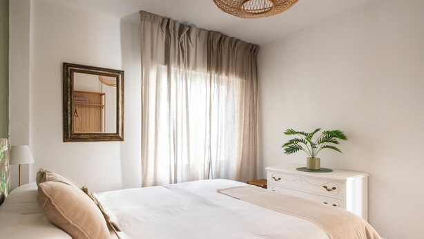 A neatly arranged bedroom with a bed, a bedside table with a plant, a mirror, and a window with curtains. - Casa Camilo – Retro con Encanto (Santander)