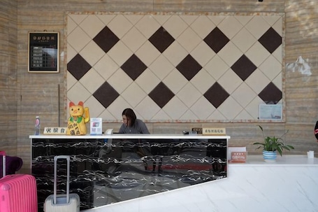 A reception desk with a patterned wall, a receptionist, and a sign with room rates.. Wangpuda Hostel