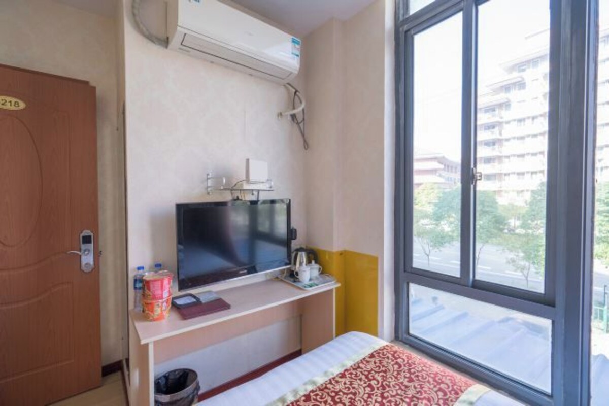 A hotel room with a bed, a TV, a desk with a computer, a window with a view of buildings, and an air conditioning unit.