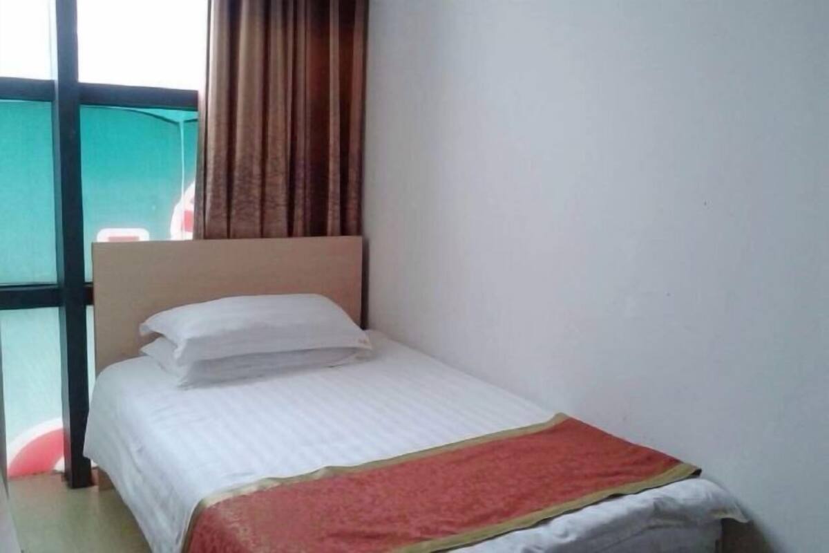 A single bed with white bedding and a red and gold blanket, a wooden headboard, and a window with curtains.