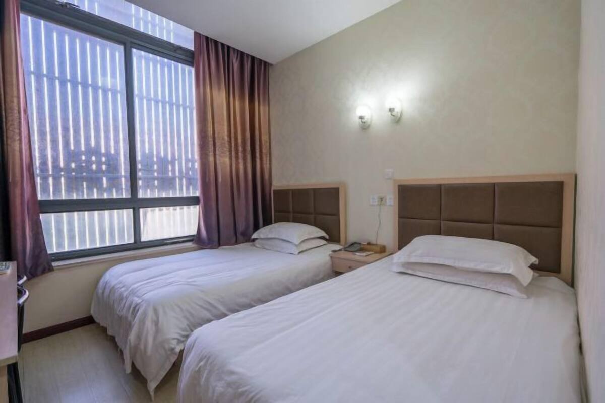 A hotel room with two beds, a window with curtains, and a wall-mounted light fixture.