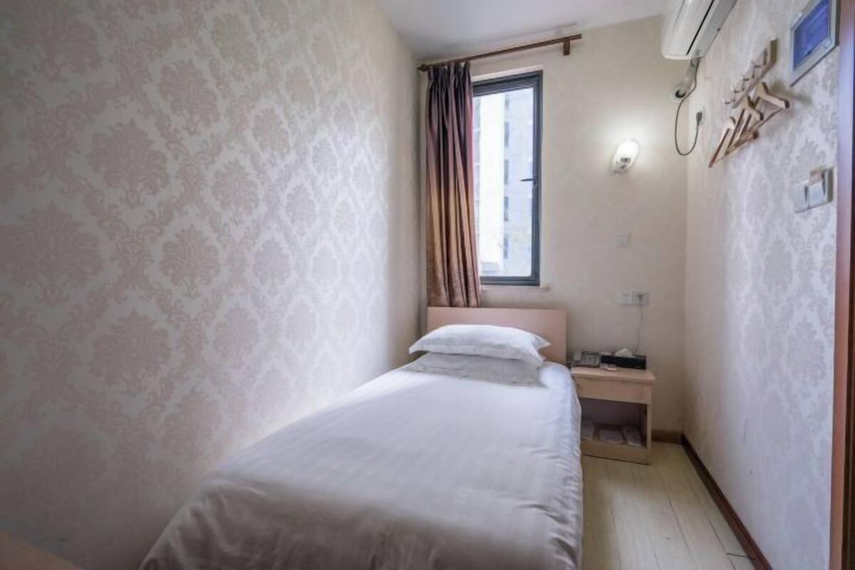 A small, single-bed room with a window, a bedside table, and an air conditioning unit.